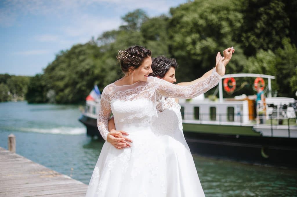 Two brides in white gowns posing together on a dock with a boat behind.