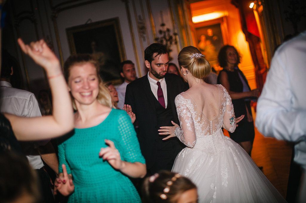 Bride in backless lace gown dances with groom surrounded by joyful guests at French chateau wedding reception.