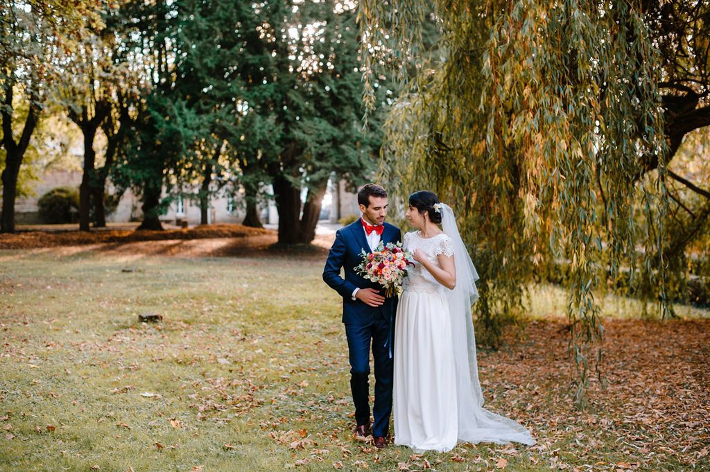 Bride and groom stand together under weeping willow tree surrounded by autumn foliage.