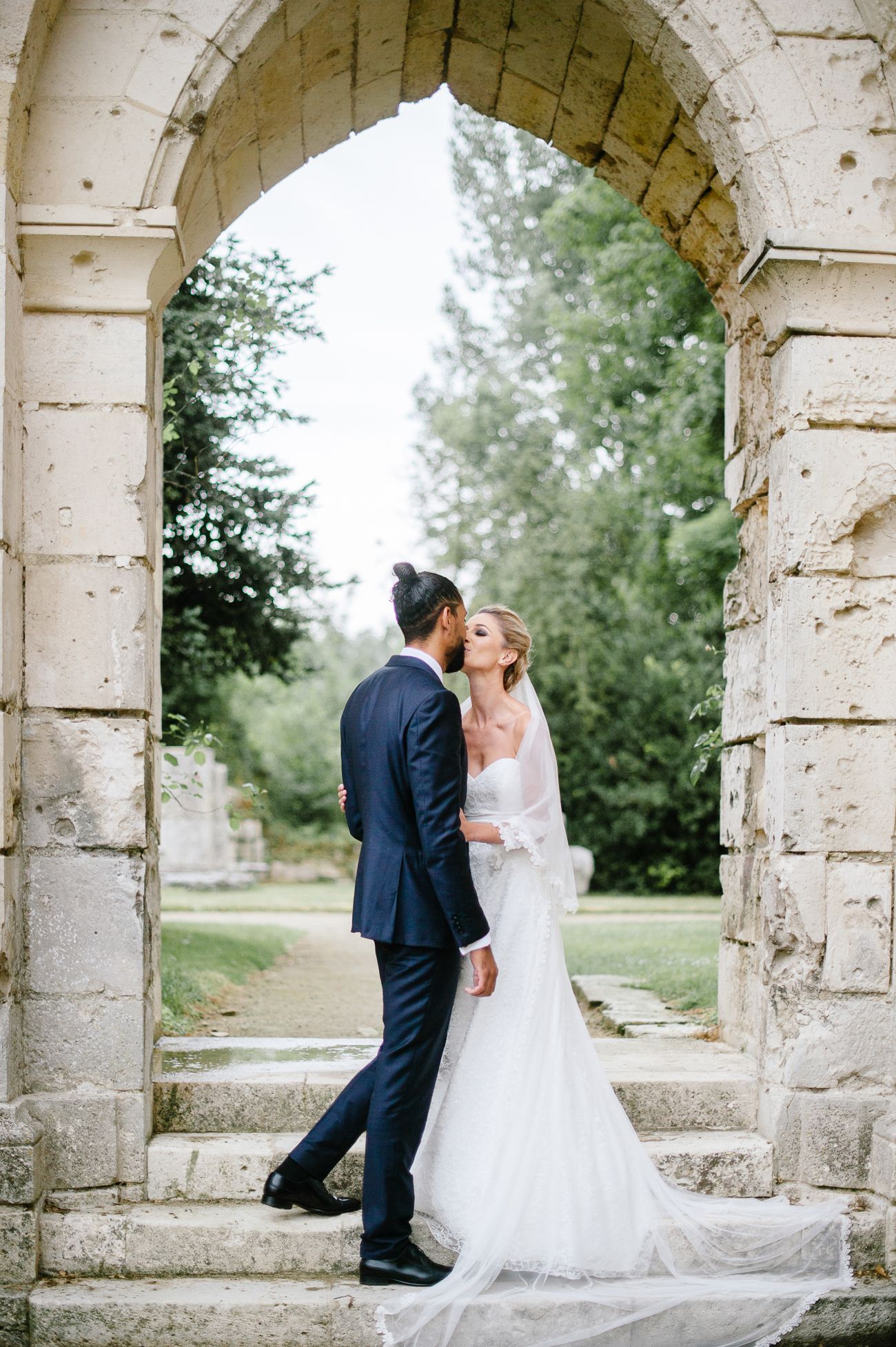 Bride and groom sharing a romantic kiss under a stone arch at an elegant outdoor wedding venue.