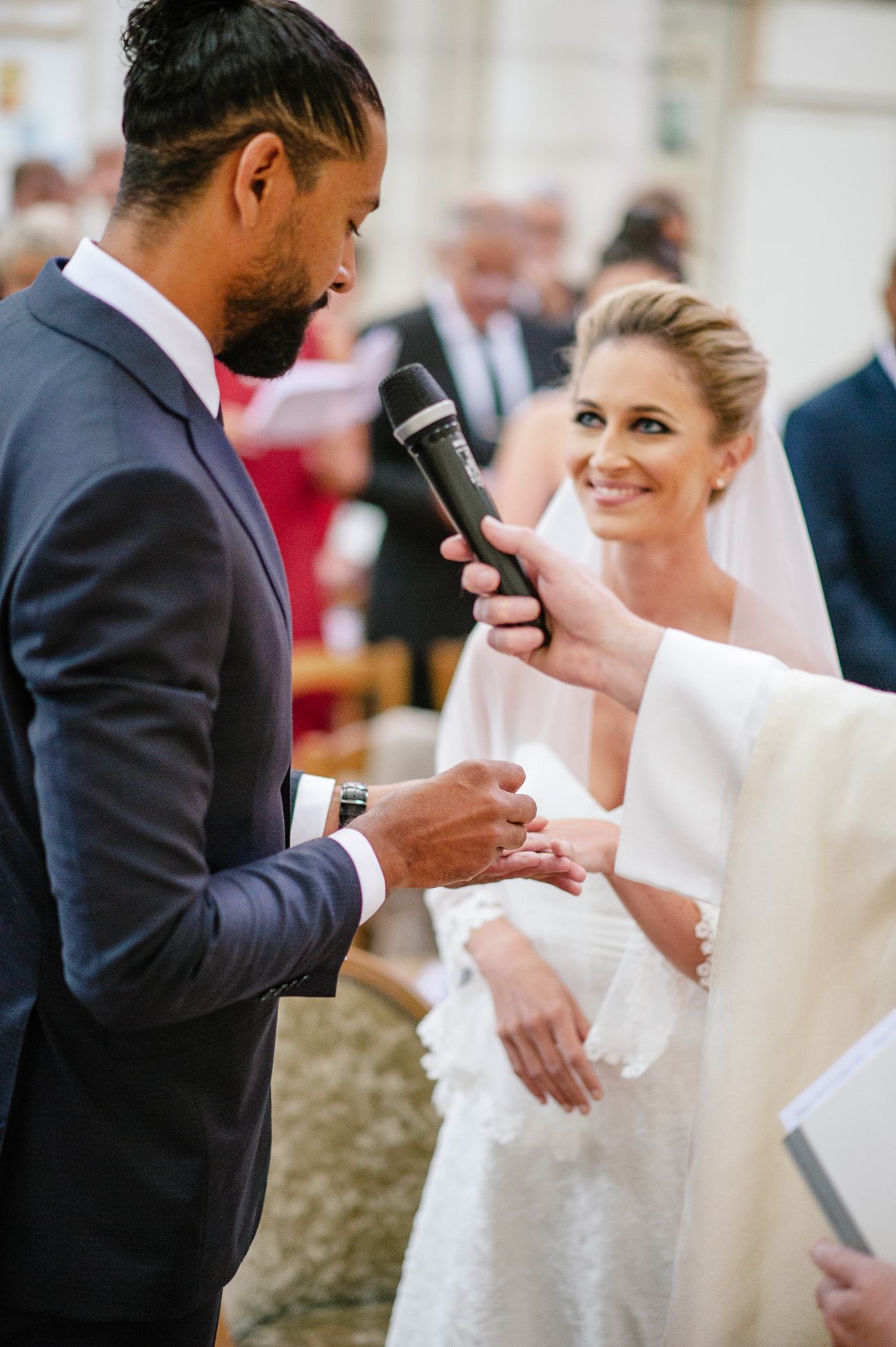 Bride smiles as groom places wedding ring on her finger during ceremony with microphone held nearby.
