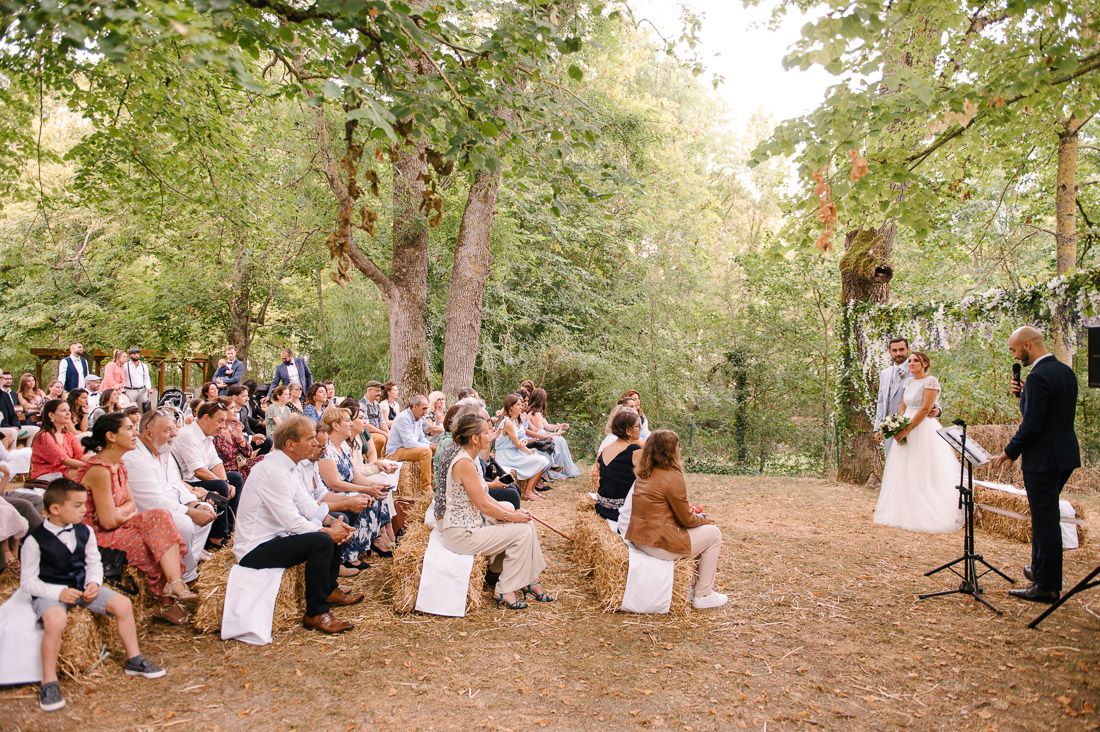 Outdoor wedding ceremony with guests seated on hay bales in a wooded forest setting.