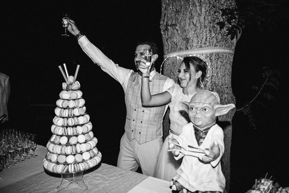 Bride and groom in masks toast beside macaron tower with Yoda figurine at wedding reception.