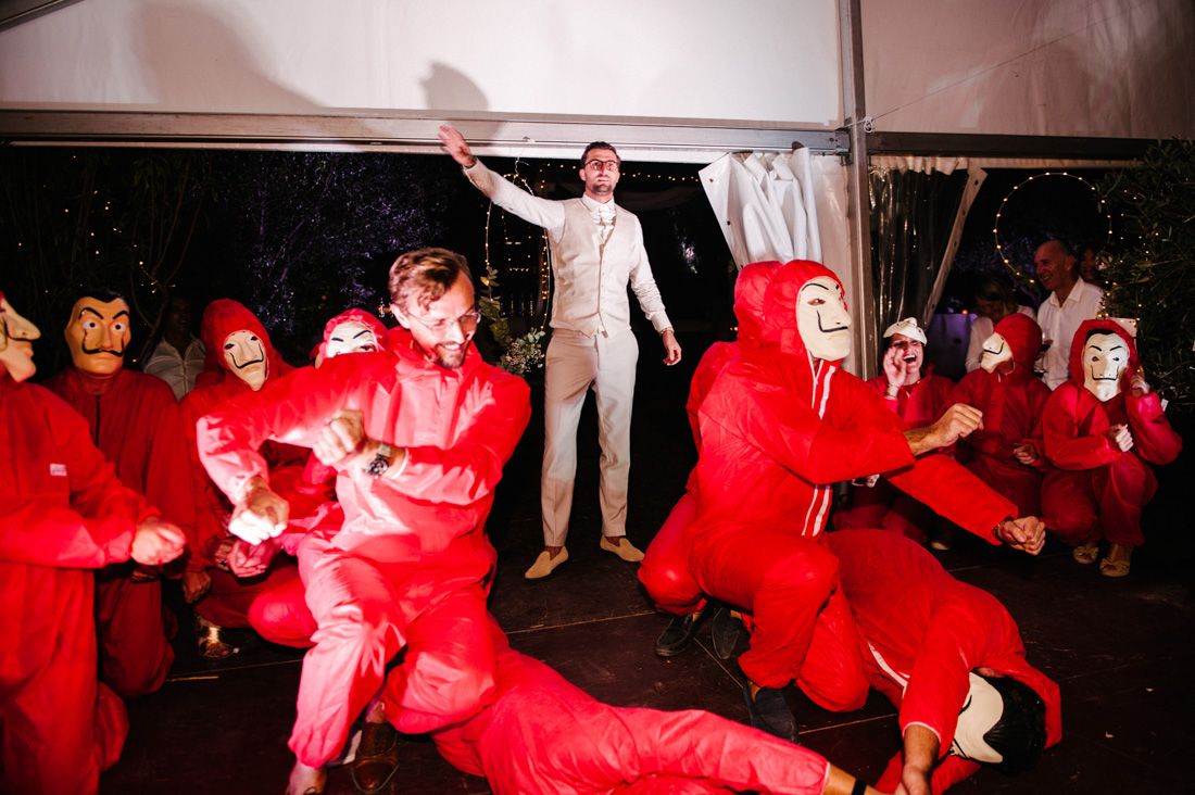 Groom in white suit surrounded by dancers in red La Casa de Papel costumes at wedding.