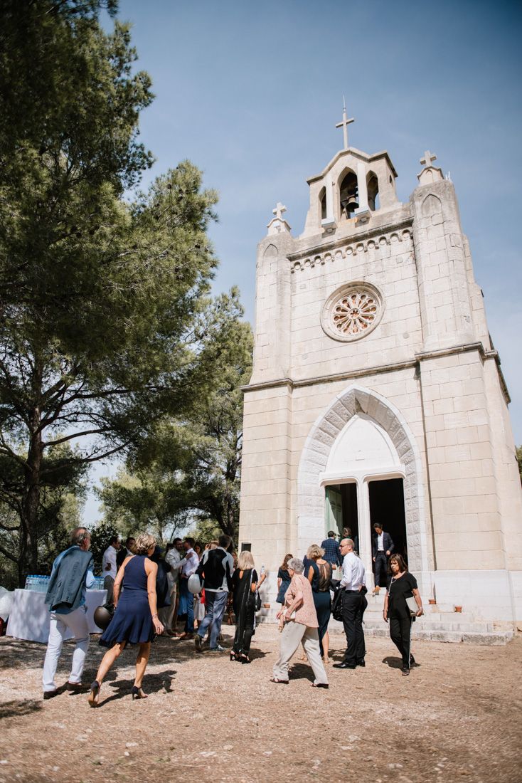 Wedding guests gather outside a small stone chapel with a cross and rose window under blue sky.