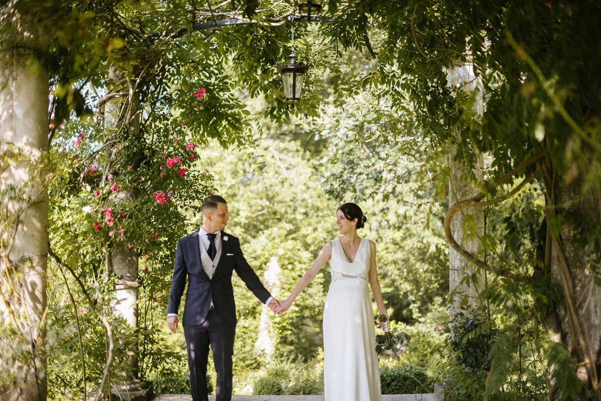 Bride and groom walking hand in hand through a romantic flower-covered garden archway.