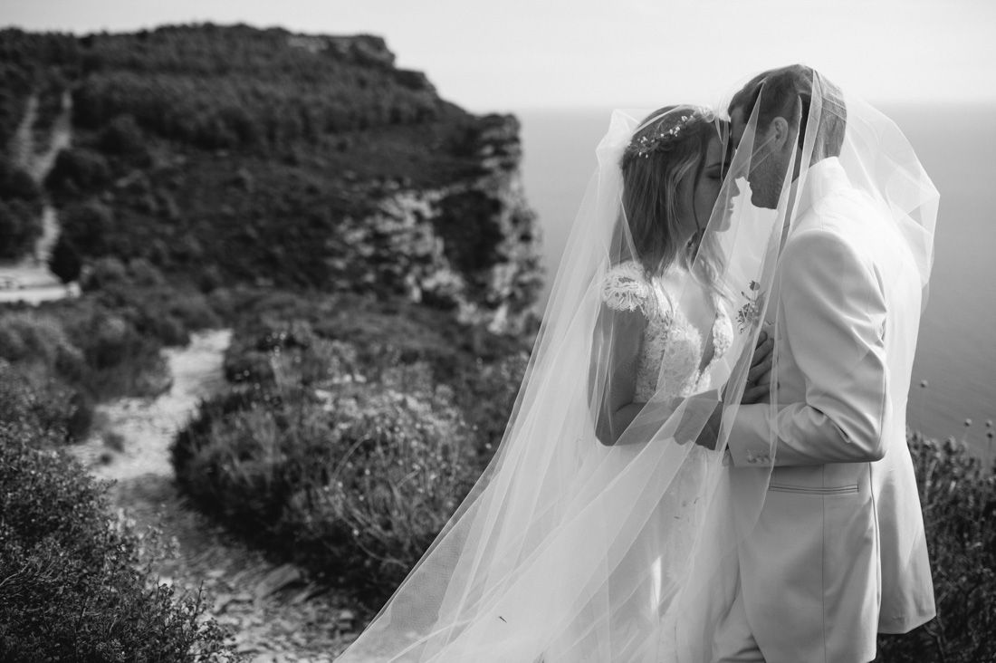Black and white wedding photo of couple kissing under veil on scenic coastal cliffside.
