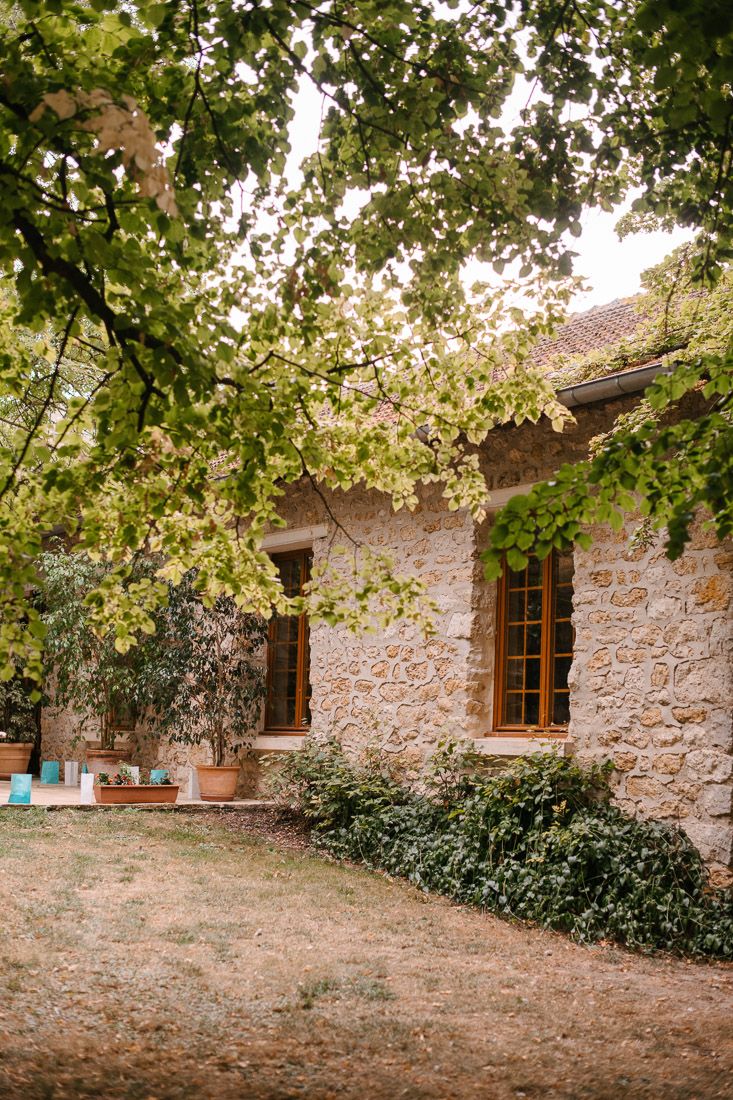 Rustic stone farmhouse with wooden windows surrounded by lush green trees and ivy in the French countryside.