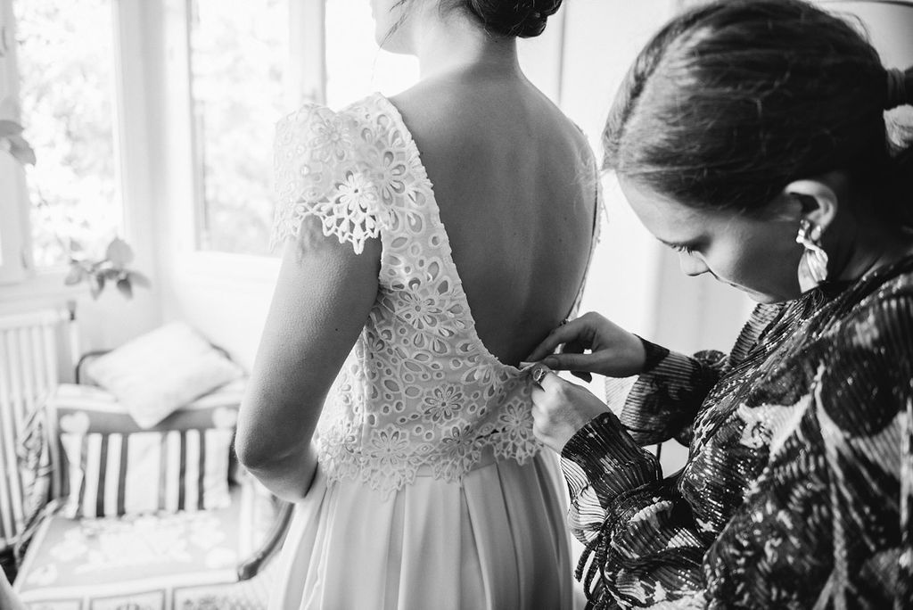 Bride in lace wedding dress being buttoned up by bridesmaid in black and white photo.