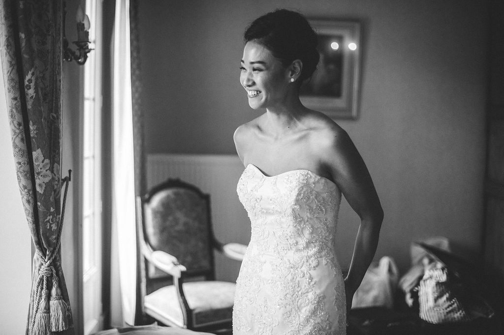 Smiling bride in strapless lace wedding gown standing by window in black and white photo.
