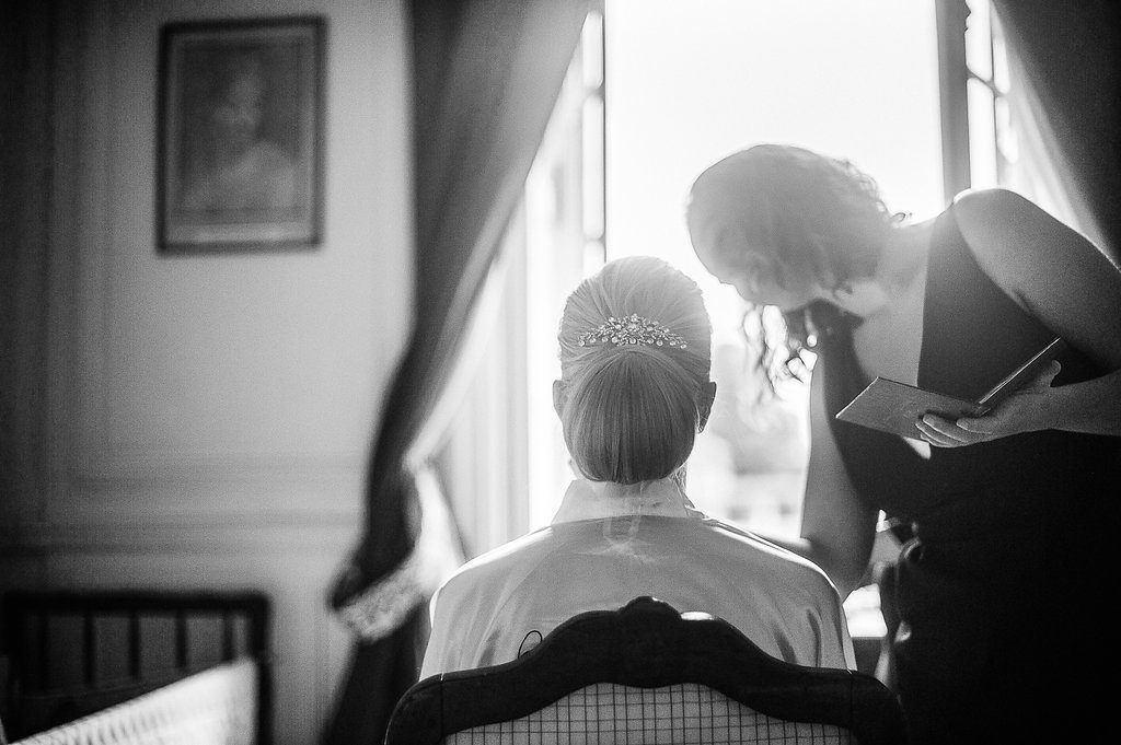 Black and white photo of bride with tiara sitting by window with wedding assistant.
