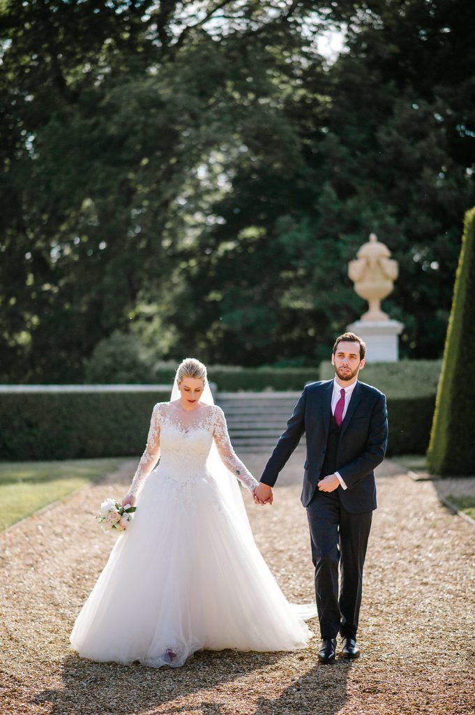 Bride and groom walking hand in hand on gravel path through elegant formal garden.