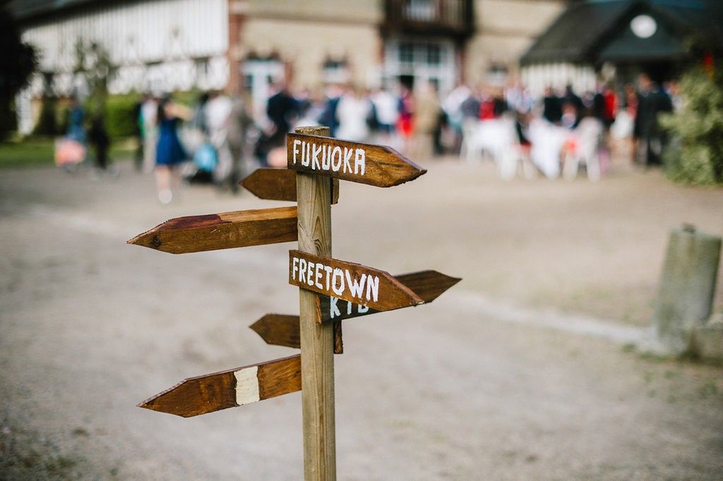 Rustic wooden directional signpost with Fukuoka and Freetown signs at an outdoor wedding venue with guests mingling.