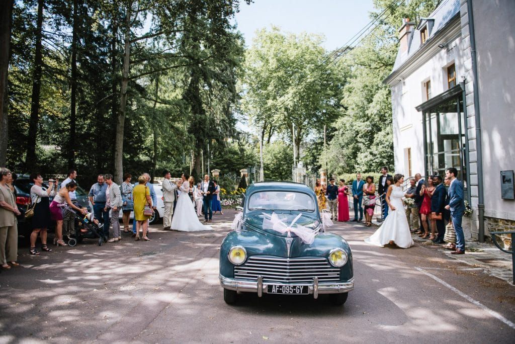 Vintage blue classic car arrives at French chateau wedding with guests gathered outside among tall trees.