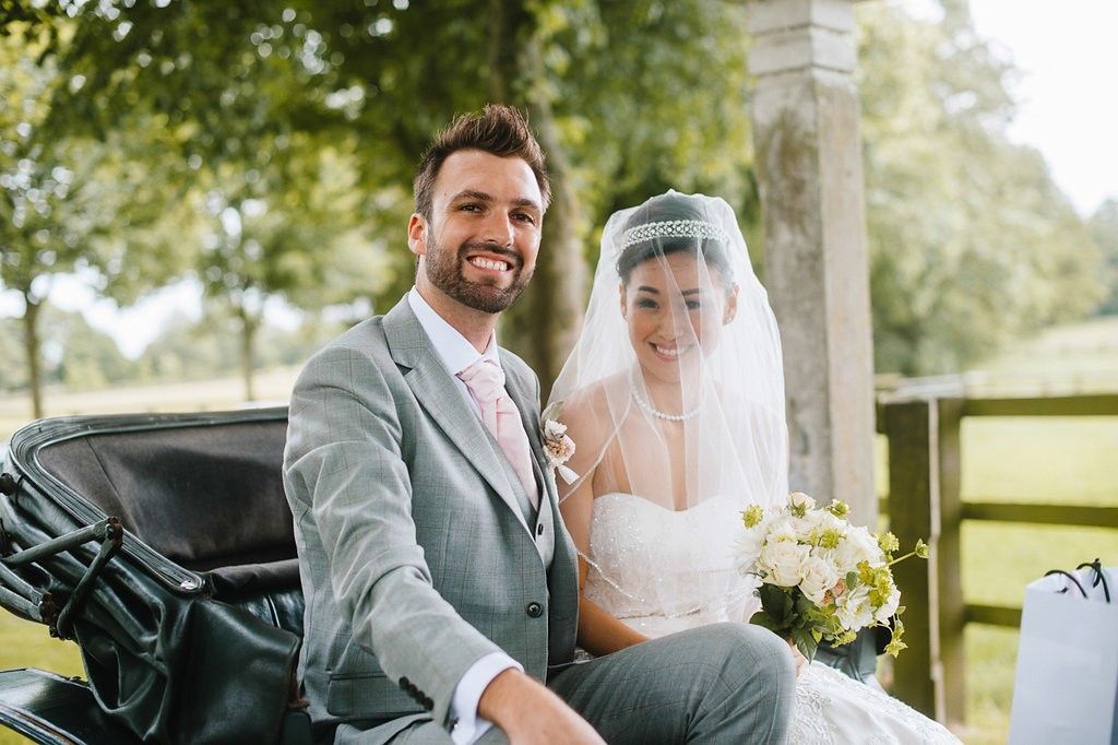 Smiling bride and groom sit together in a horse-drawn carriage, bride wearing veil and holding bouquet.