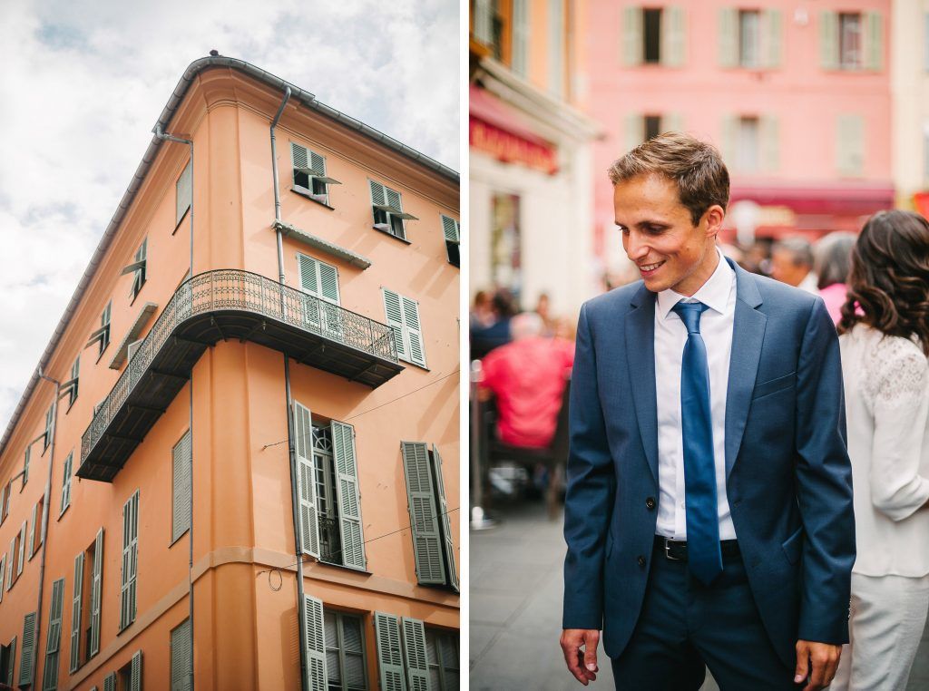Dapper groom in blue suit smiles beside colorful Mediterranean architecture in Nice, France on wedding day.