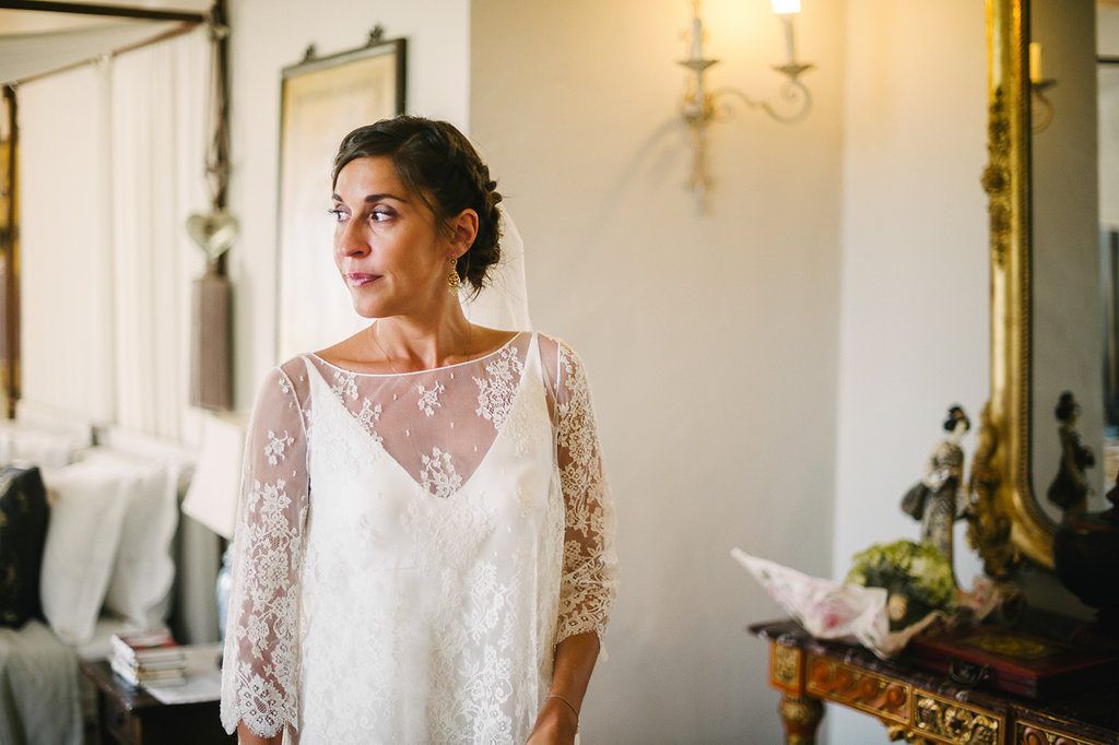 Bride in lace long-sleeve wedding dress standing by ornate gold mirror in sunlit room.