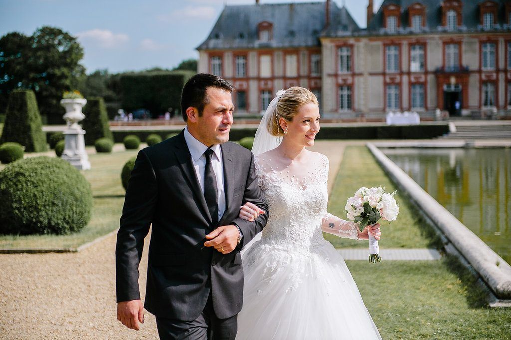 Bride and groom walk arm in arm through formal French château garden with manicured topiary and reflecting pool.