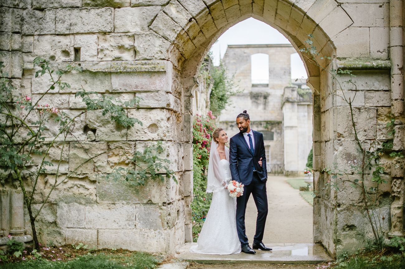 Elegant bride and groom stand beneath weathered stone archway at rustic outdoor wedding venue.