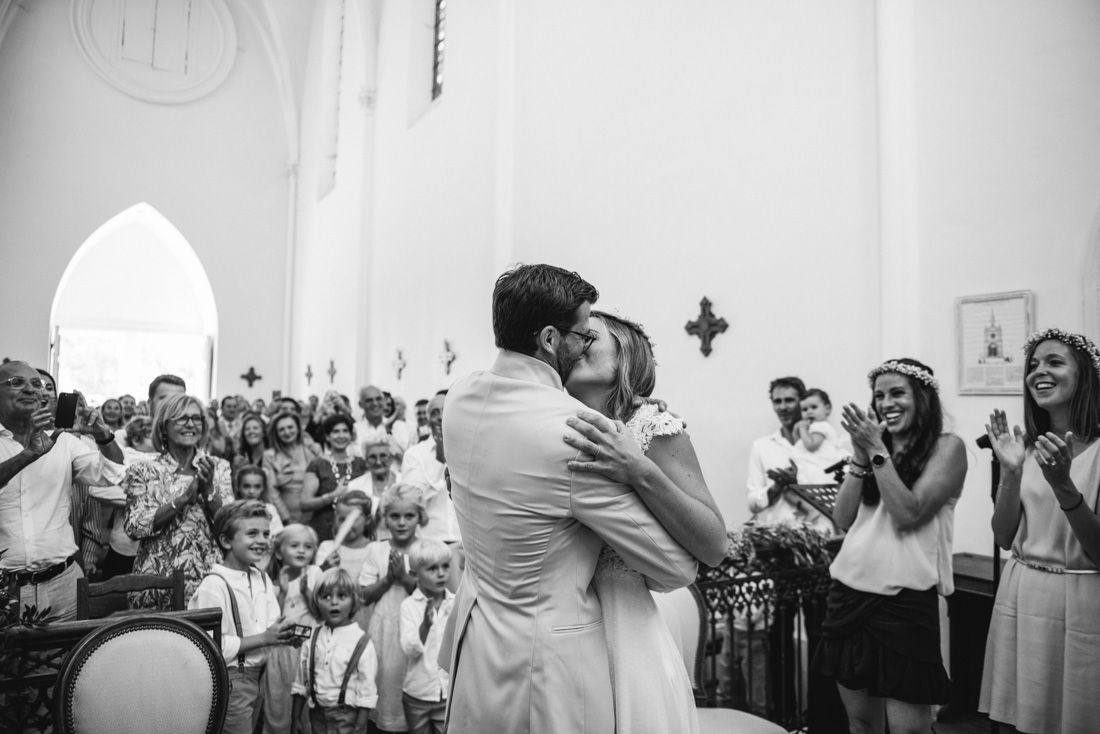 Bride and groom share first kiss at altar surrounded by cheering wedding guests.
