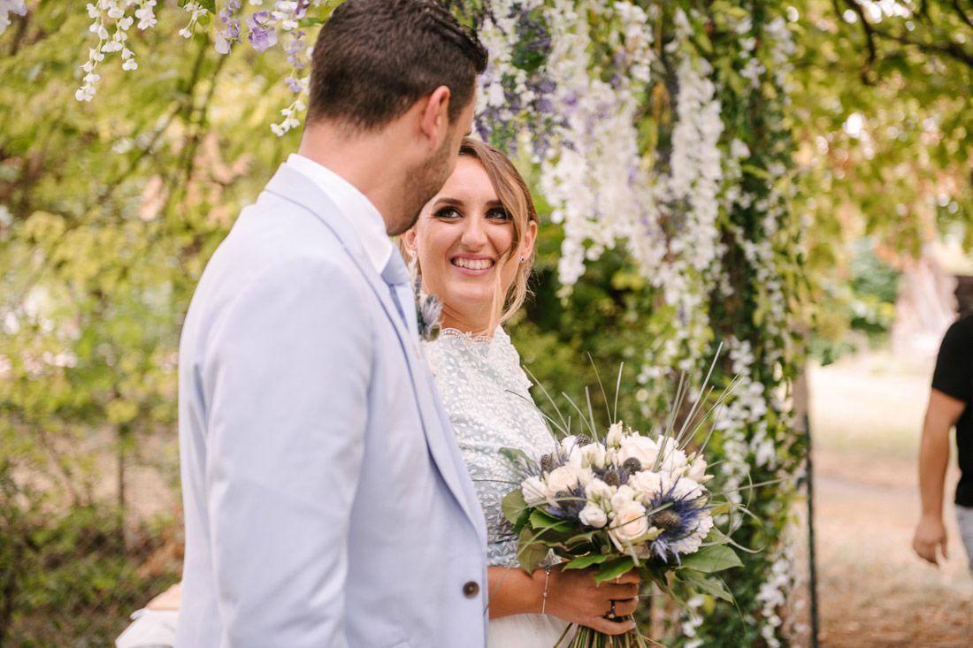 Bride smiling holding a bouquet of white and blue flowers with groom under a floral arch outdoors.