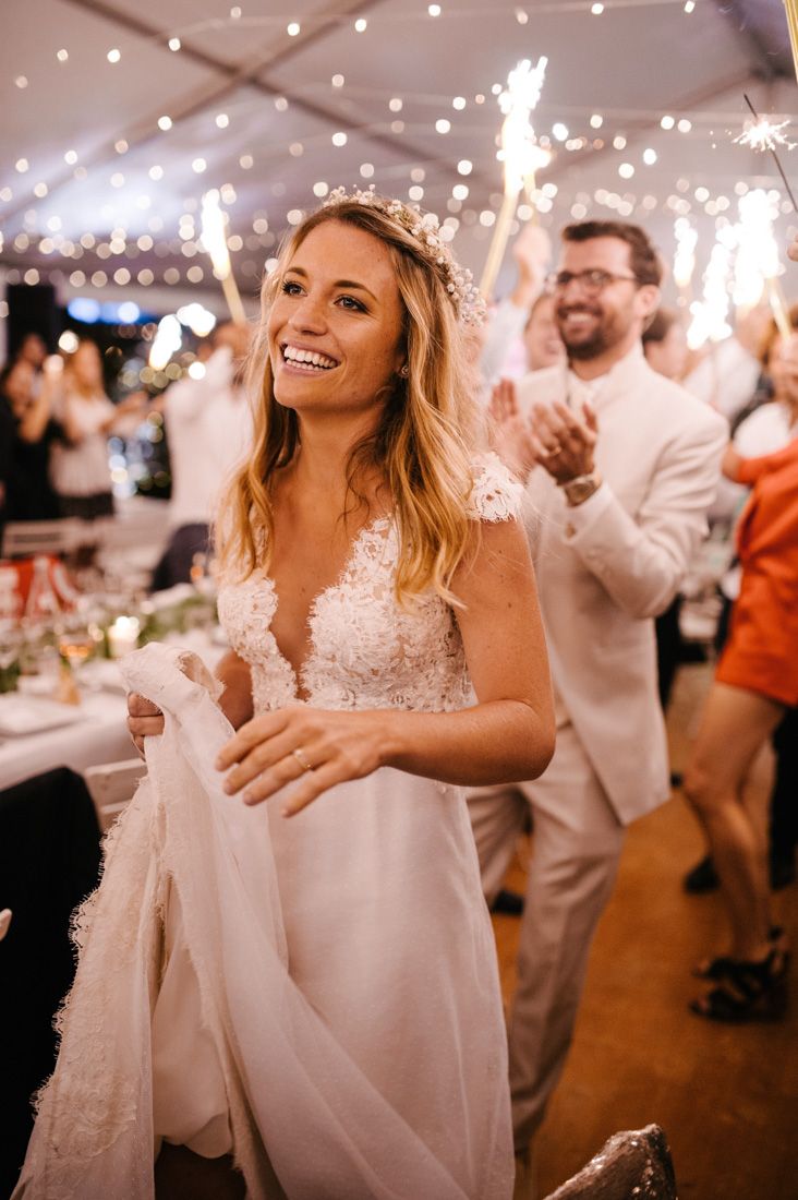 Radiant bride in lace gown smiling under fairy lights with sparklers at wedding reception.