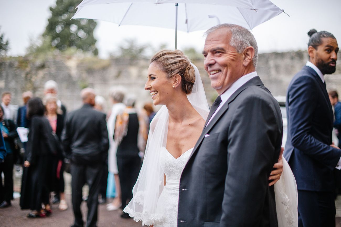 Bride in white gown and veil walks with older gentleman under umbrella at outdoor wedding venue.