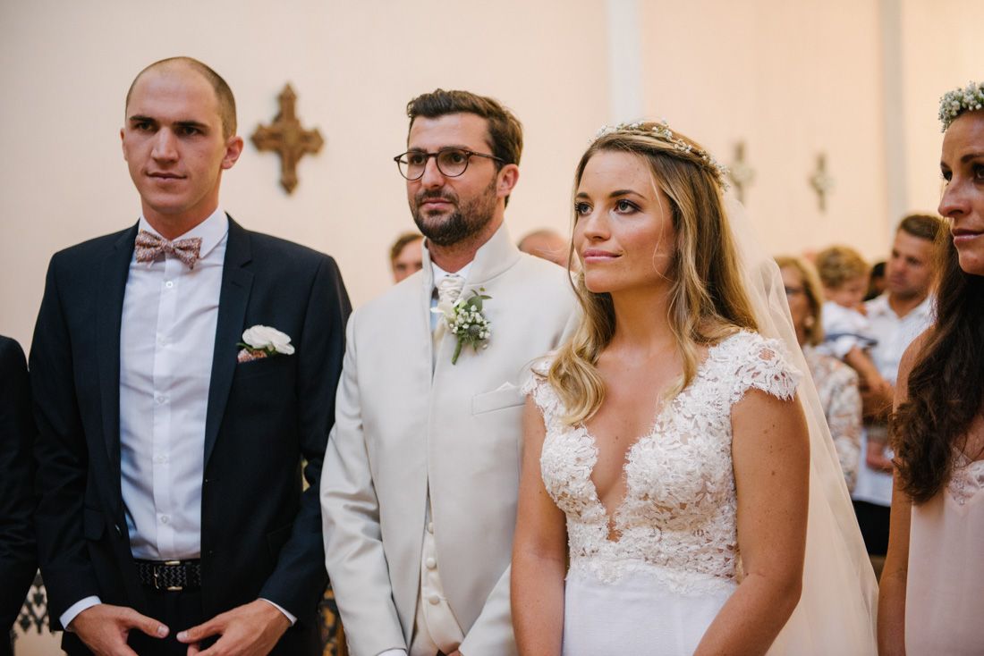 Bride in lace gown stands with groom in white suit and groomsman during church wedding ceremony.