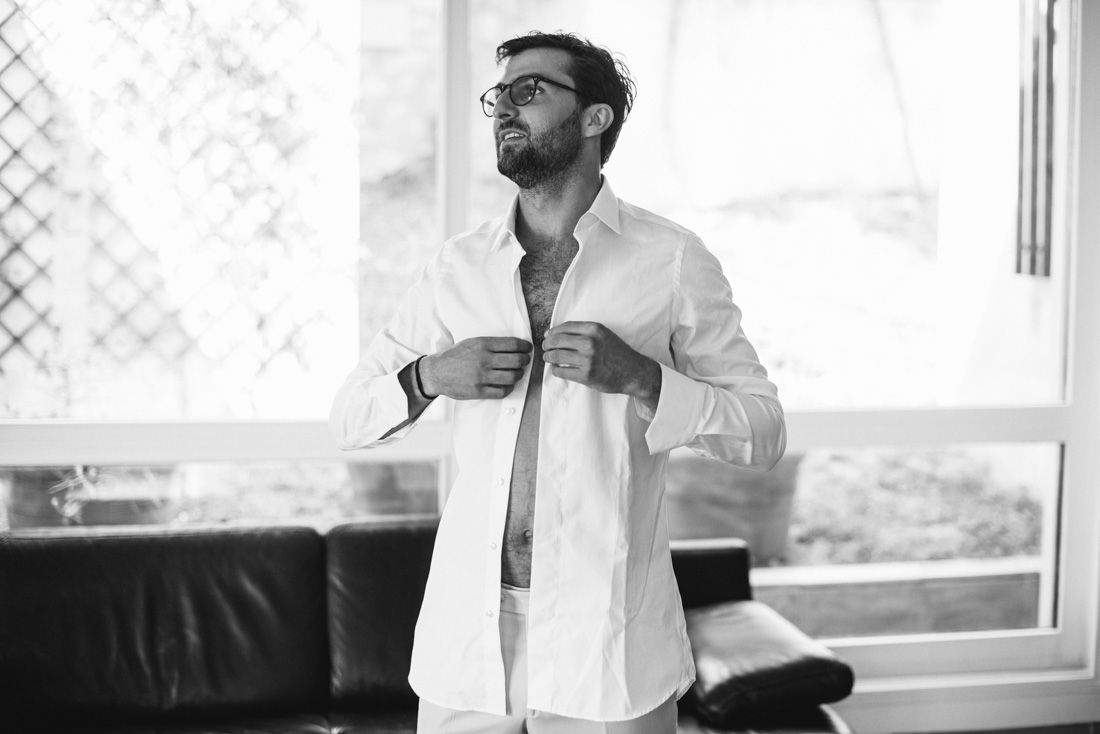 Black and white photo of a man in glasses buttoning a white dress shirt near a window.