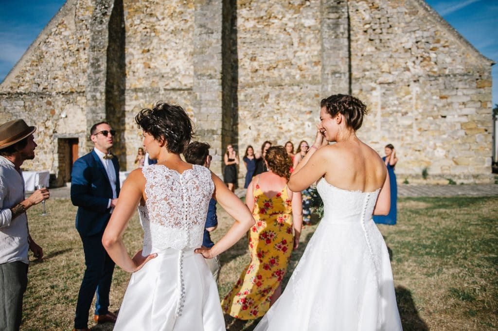 Two brides in white wedding gowns walk together outside a rustic stone barn with guests.