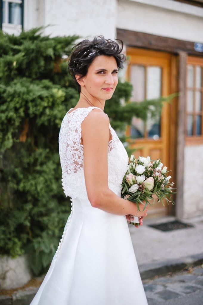 A bride with short dark hair in a sleeveless lace wedding dress holds a bouquet of white and pink flowers outdoors.