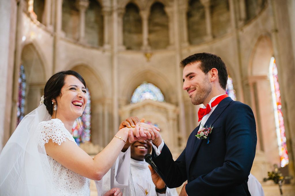 Laughing bride and smiling groom exchange rings during joyful church wedding ceremony with colorful stained glass.
