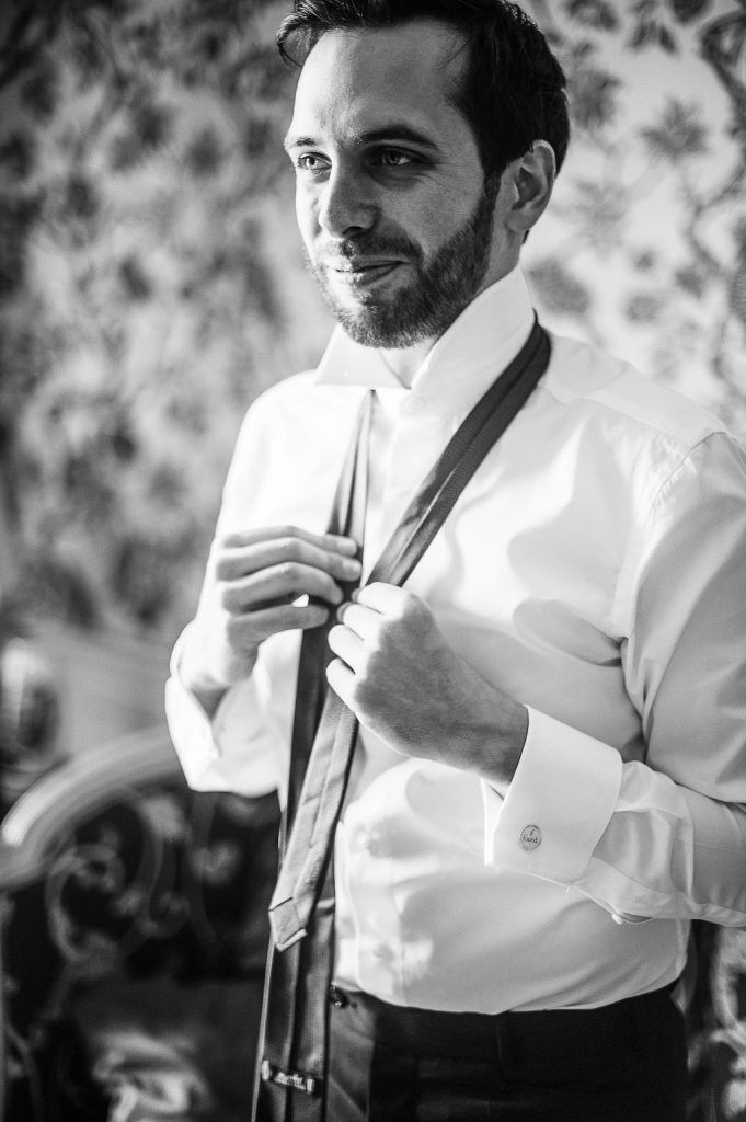 Groom in white dress shirt adjusting tie in black and white wedding preparation portrait.