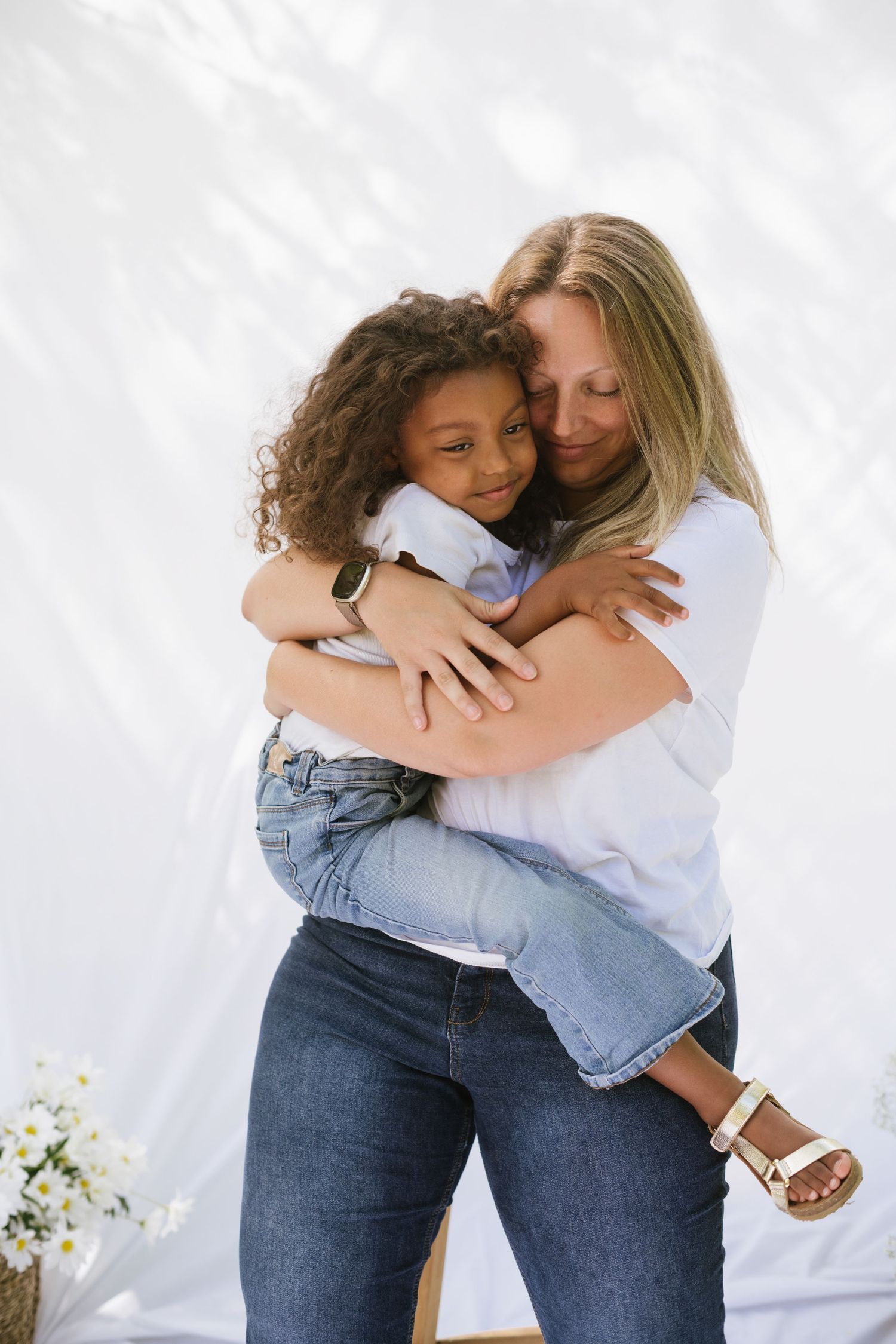 A blonde mother embraces her curly-haired daughter lovingly in front of a white fabric backdrop.