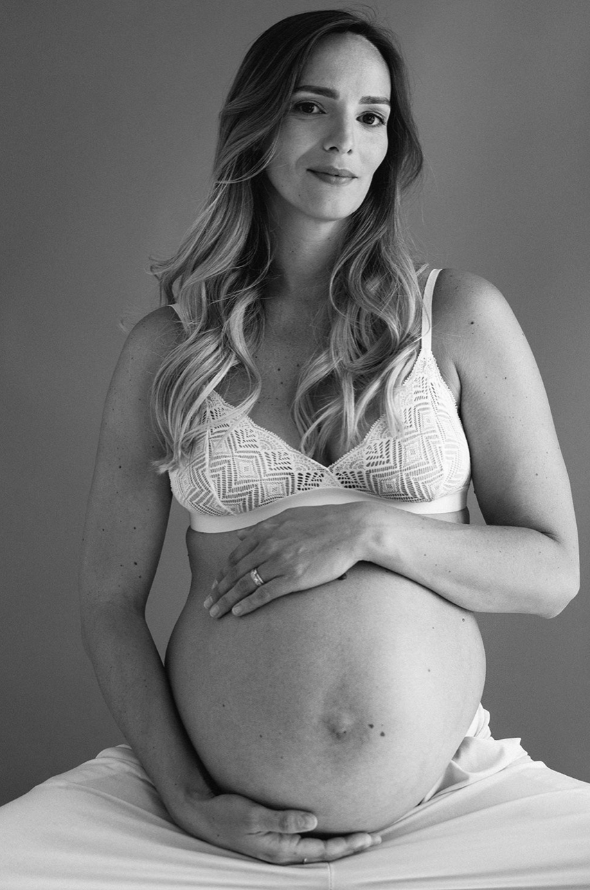 Black and white photo of pregnant woman in lace bra holding her bare baby bump while seated.