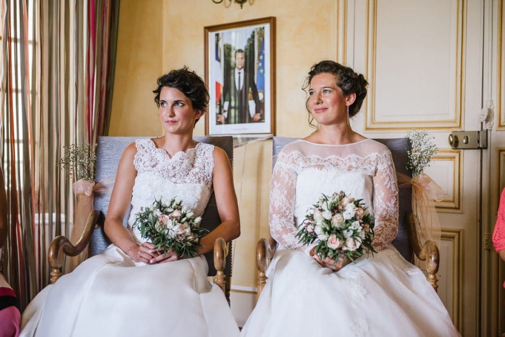 Two brides in matching white lace wedding gowns holding bouquets during civil ceremony at French town hall.