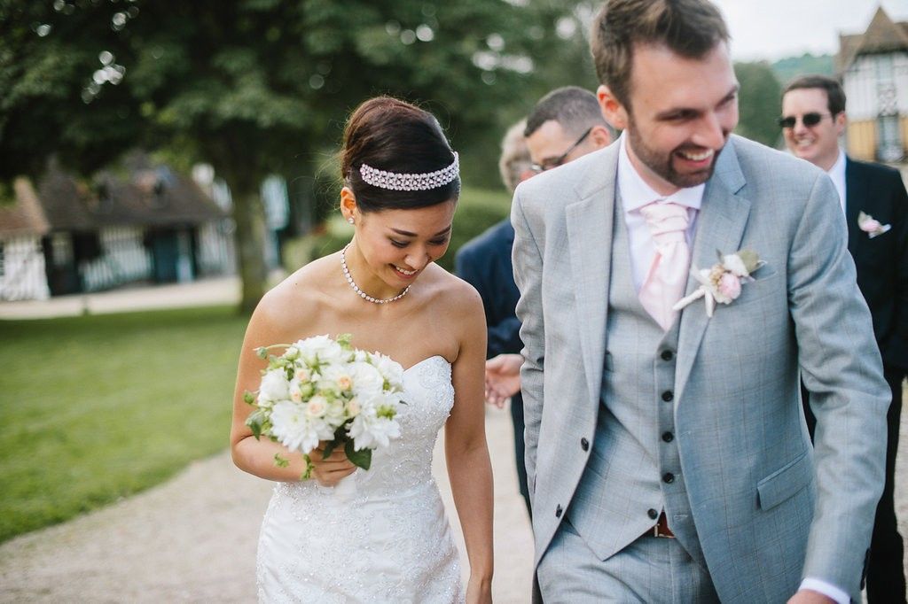Elegant bride in strapless gown and crystal headband walks arm in arm with smiling groomsman holding white bouquet.
