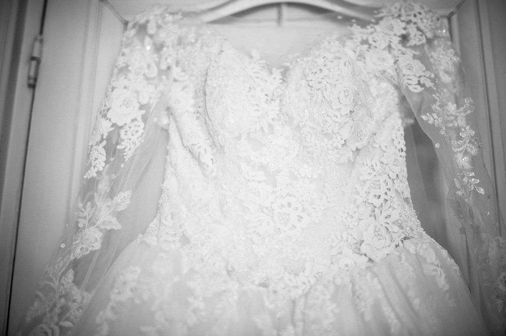 Black and white close-up of intricate lace and embroidered wedding dress hanging on wardrobe.