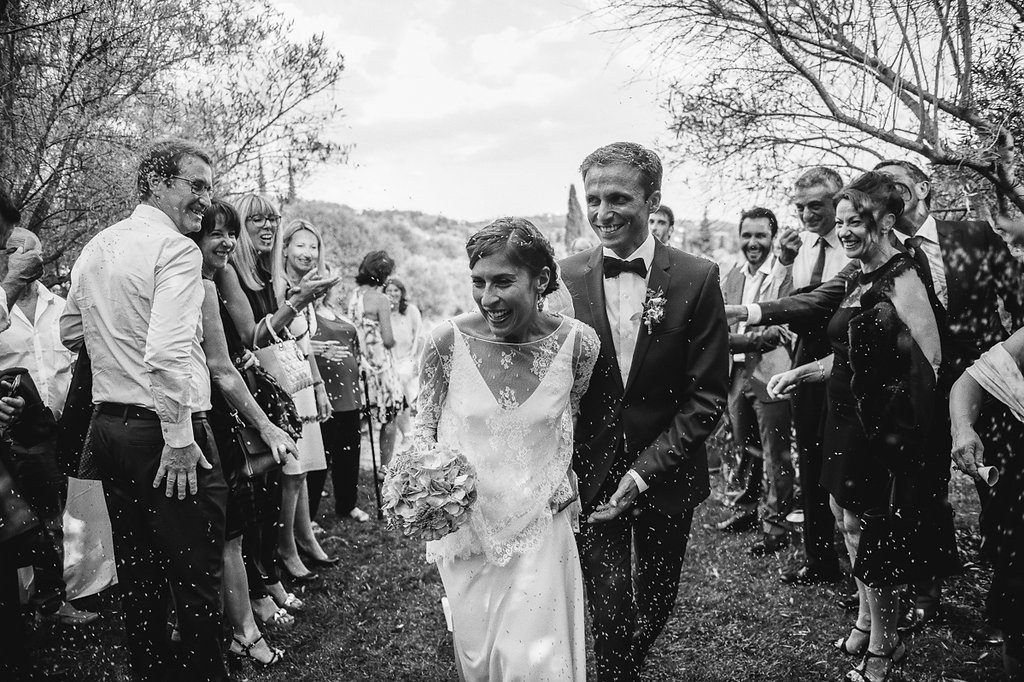 Black and white photo of newlyweds walking through confetti shower surrounded by cheering wedding guests outdoors.