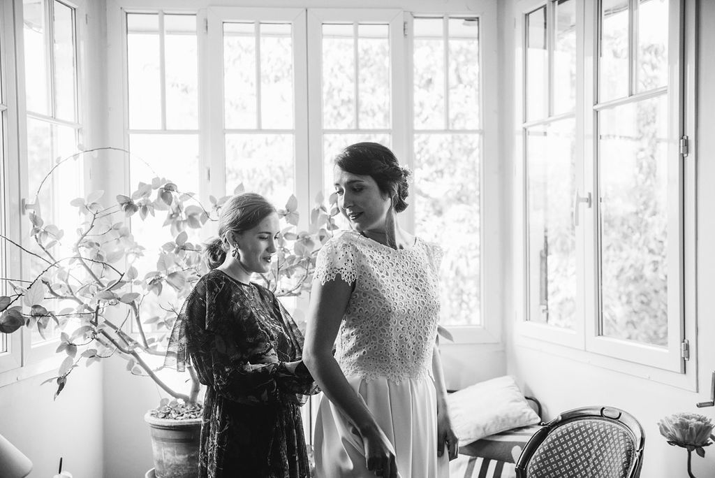 Bride in elegant white gown standing by window with helper in black and white wedding photo.
