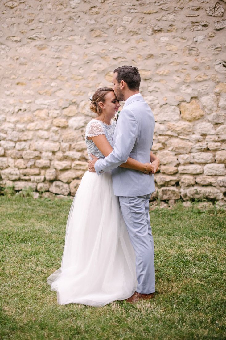 Bride and groom embrace romantically in front of rustic stone wall on grass.