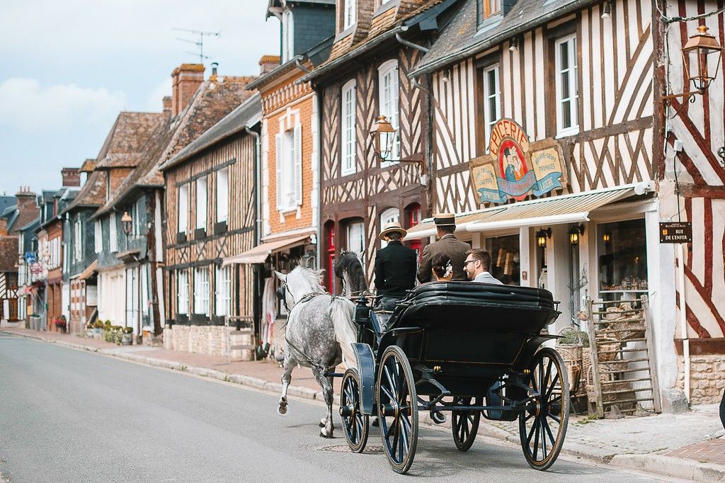 Horse-drawn carriage rides through a picturesque French village street lined with colorful timber-framed buildings.