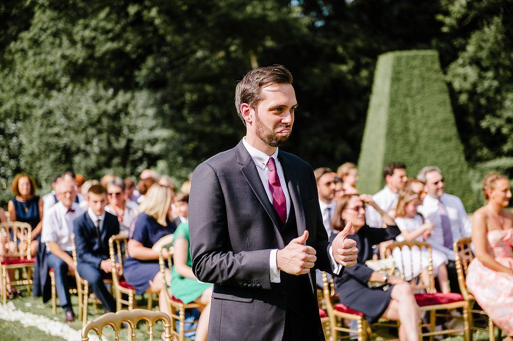 Smiling groomsman gives thumbs up at outdoor wedding ceremony with seated guests in garden setting.
