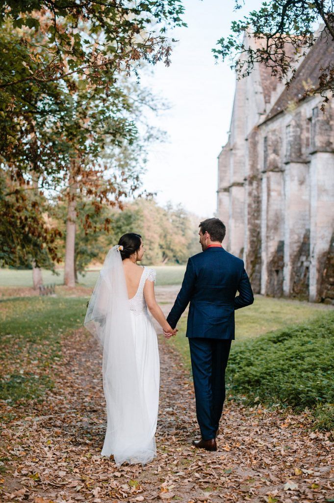 Bride and groom walk hand in hand along leaf-covered path beside historic stone church.