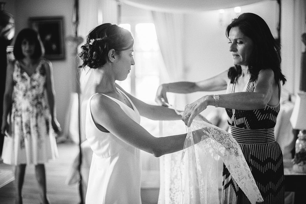 Black and white photo of bride being helped into wedding dress by two women in elegant room.