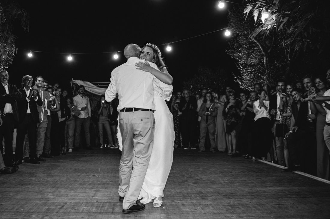 Black and white photo of bride and groom sharing first dance under string lights outdoors.