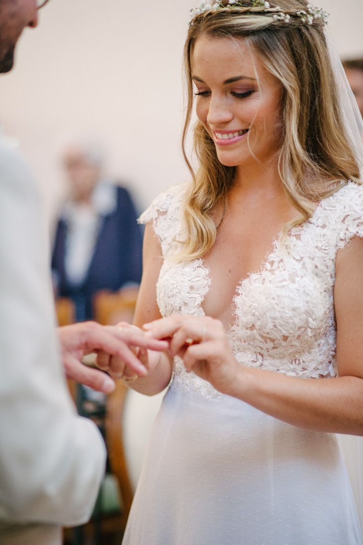 Smiling bride in lace gown and floral crown exchanges wedding rings at altar.