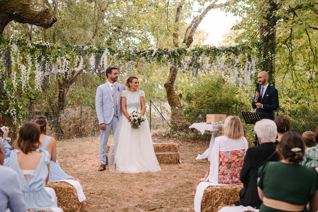 Bride and groom stand together during outdoor rustic woodland wedding ceremony with floral garland arch and hay bale seating.