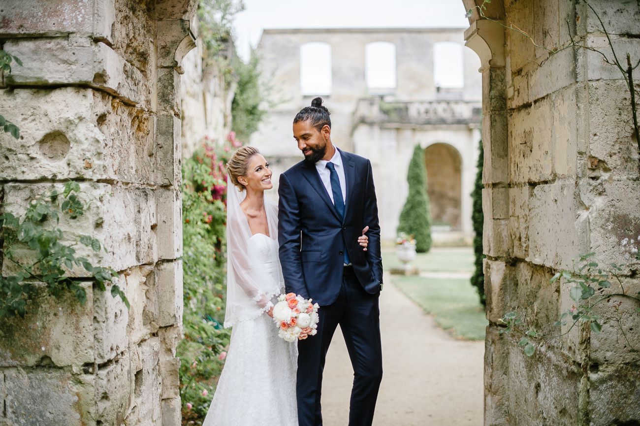 Newlyweds share romantic moment between ancient stone columns, groom in navy suit, bride with floral bouquet.