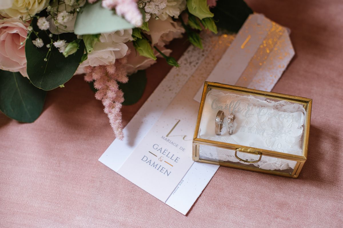 Wedding rings in a gold glass box beside an invitation card and floral arrangement.